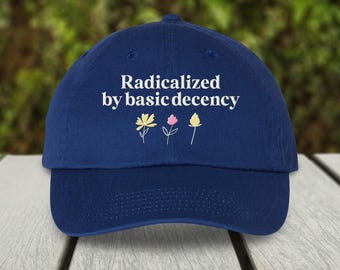 May include: A navy blue baseball cap with the phrase "Radicalized by basic decency" in white lettering. Below the text are three small, simple flower illustrations in yellow, pink, and yellow. The cap is sitting on a white wooden surface.