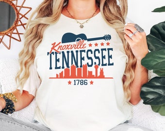 May include: White t-shirt with a blue and red graphic design featuring a guitar, the text "Knoxville Tennessee", a skyline, and the year "1786".