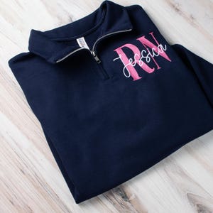 May include: A navy blue quarter-zip sweatshirt with the letters "RN" embroidered in pink and the name "Jessica" embroidered in white below.