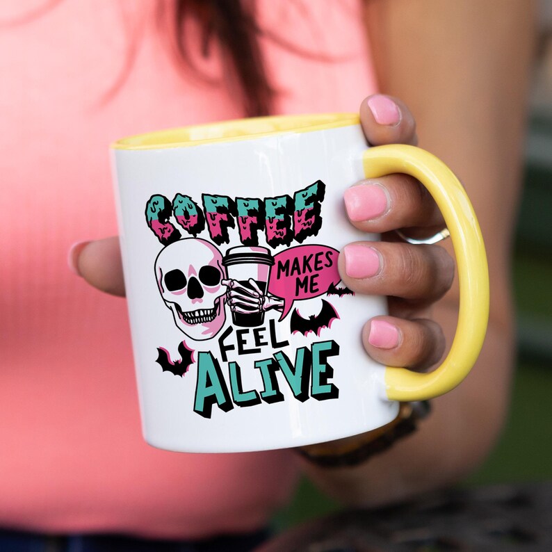 May include: White ceramic coffee mug with a yellow handle and interior. The mug features a skull graphic holding a coffee cup, with the words "COFFEE MAKES ME FEEL ALIVE" in a spooky font. The design includes bats and pink and teal accents.