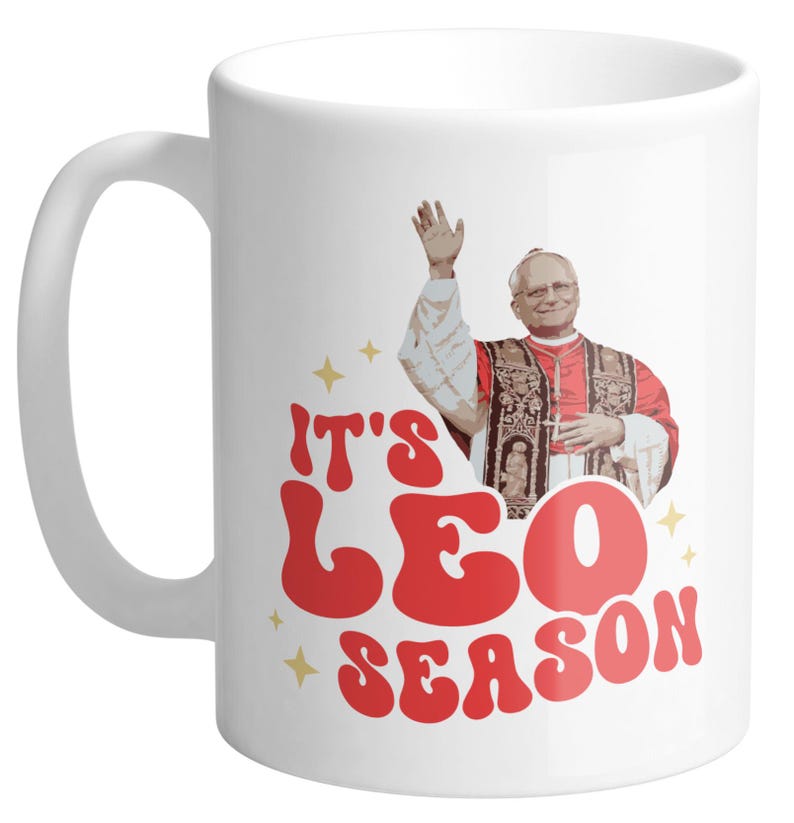 May include: White ceramic mug with a red and white graphic that says "It's Leo Season". The graphic features a cartoon image of a man in a red robe and a white hat.