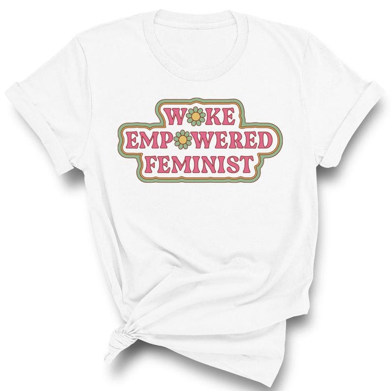 May include: White t-shirt with the text "WOKE EMPOWERED FEMINIST" in a retro-style design. The text is in pink, green, and yellow, with flower accents. The shirt is folded and knotted at the bottom.