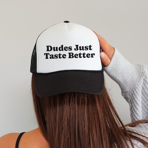May include: A white and black trucker hat with the text "Dudes Just Taste Better" in bold, black letters. The hat has a black brim and mesh back. The hat is being worn by a person with long brown hair.