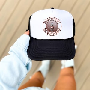 May include: Black and white trucker hat with a white front panel. The front panel features a circular graphic with the text "Pepper University Doctorate Program" around a soda bottle illustration with the number "23".