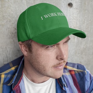 May include: A bright green baseball cap featuring the white embroidered text I WORK HERE centered on the front panel. The cap has a structured crown, a curved brim, and a fabric strap closure at the back, providing a classic athletic look.