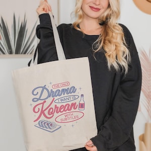 May include: Beige tote bag with the text "The Only Drama I Want Is Korean With Subtitles" in blue and pink. The design includes a bottle and sushi illustration. The bag has gray straps.