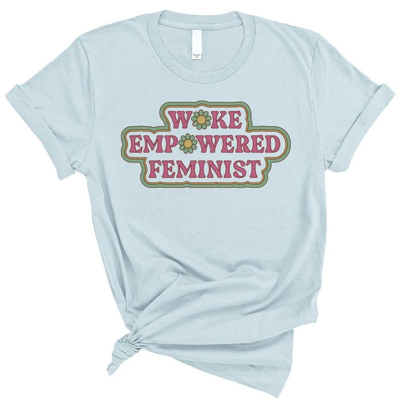 May include: Light blue t-shirt with rolled-up sleeves. The shirt features the text "WOKE EMPOWERED FEMINIST" in a retro-style design with flowers. The text is in a pink and green color scheme.