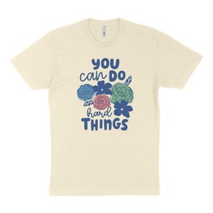 Puede incluir: Camiseta color crema con el texto "YOU can DO hard THINGS" en azul. El dise&ntilde;o incluye un grupo de flores azules, verdes y rosas. La camiseta tiene cuello redondo y mangas cortas.