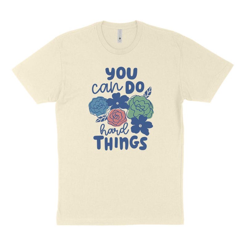 Puede incluir: Camiseta color crema con el texto "YOU can DO hard THINGS" en azul. El dise&ntilde;o incluye un grupo de flores azules, verdes y rosas. La camiseta tiene cuello redondo y mangas cortas.