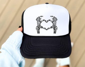 May include: Black and white trucker hat featuring a heart made of skeleton hands on the front. The hat has a white front panel and a black brim and mesh back. The hat is being held up by a person.