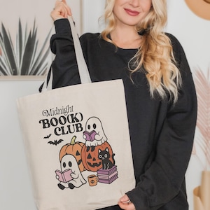 May include: Beige tote bag with the text "Midnight BOO(K) CLUB" and Halloween-themed illustrations. The design includes ghosts reading books, pumpkins, a black cat, and bats. The bag has gray straps.