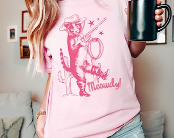 May include: A light pink t-shirt featuring a pink illustration of a cat dressed as a cowboy, holding a rifle and lasso, with the word "Meowdy!" below. The cat is wearing a cowboy hat and boots. The shirt is being held by a person.