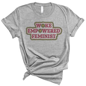 May include: A gray t-shirt with the words "WOKE EMPOWERED FEMINIST" in a retro-style design. The text is in a pink and green color scheme, with a flower accent. The shirt is knotted at the bottom.
