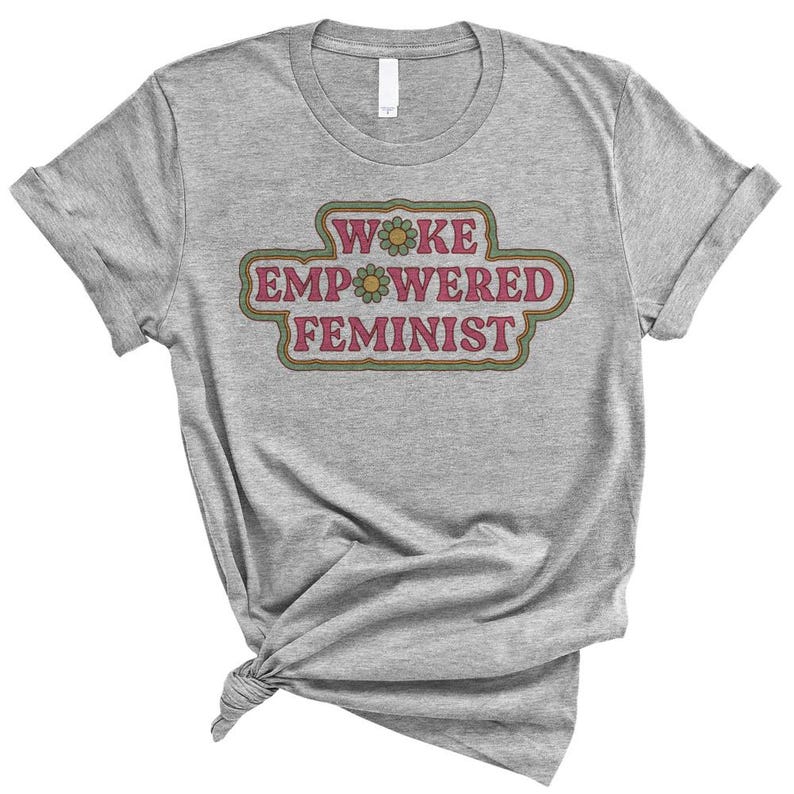 May include: A gray t-shirt with the words "WOKE EMPOWERED FEMINIST" in a retro-style design. The text is in a pink and green color scheme, with a flower accent. The shirt is knotted at the bottom.