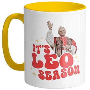 May include: White ceramic mug with a yellow rim. The mug features a retro-style graphic of a man in a red robe with the text "It's Leo Season".