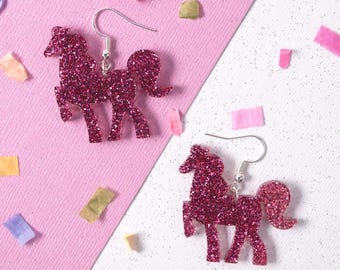 May include: A pair of pink glitter unicorn earrings. The earrings feature a detailed unicorn silhouette with a hook closure. The earrings are set against a pink and white background with confetti.