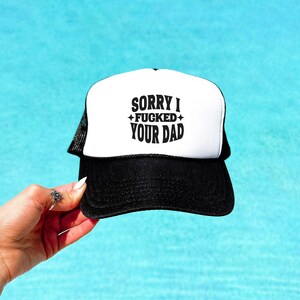 May include: A black and white trucker hat featuring a white front panel with the black text SORRY I FUCKED YOUR DAD printed in a bold font, accompanied by small star icons. The hat has a black brim and a black mesh back panel for ventilation.