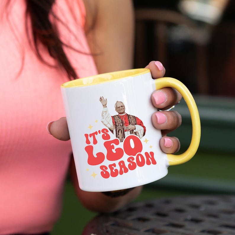 May include: White ceramic mug with a yellow rim. The mug features a graphic of a man in a red robe with the text "It's Leo Season" in red and yellow letters.