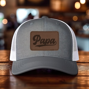 May include: A gray trucker hat with a white mesh back. The hat features a brown leather patch with the word "Papa" in a script font. The hat is sitting on a wooden surface.