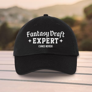 May include: A black baseball cap with the text "Fantasy Draft EXPERT" in white, with the phrase "(SINCE NEVER)" below. The cap has a curved brim and a rounded crown, suitable for casual wear.