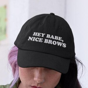 May include: Black baseball cap with white embroidered text that reads "Hey Babe, Nice Brows".