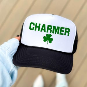 May include: A black and white trucker hat with a white front panel. The front panel has the word "CHARMER" in green letters and a green shamrock.