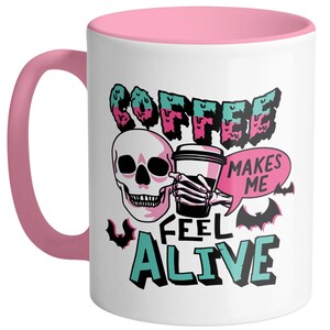 May include: White ceramic coffee mug with a pink interior and handle. The mug features a skull graphic holding a coffee cup, with the words "COFFEE MAKES ME FEEL ALIVE" in a spooky font. Bats are also included in the design.