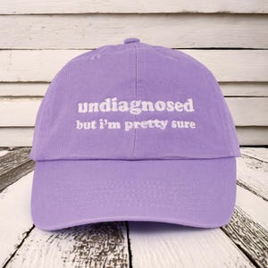 May include: Purple baseball cap with white embroidered text that reads "undiagnosed but I'm pretty sure".