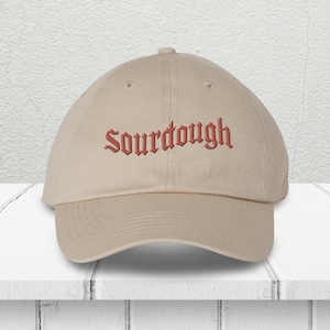 May include: A beige baseball cap with the word "Sourdough" embroidered in a reddish-orange color across the front. The cap has a curved brim and a button on top. The hat is sitting on a white surface.