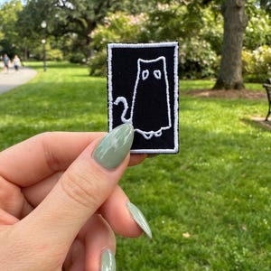 May include: A rectangular embroidered patch featuring a black background with a white stitched outline of a cat figure. The cat design includes two simple eye shapes and a curved tail, all bordered by a thin white rectangular frame.