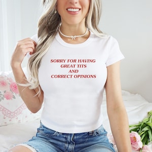 May include: White t-shirt with the text "SORRY FOR HAVING GREAT TITS AND CORRECT OPINIONS" in red. The shirt is a crew neck style and is paired with blue denim shorts. The model is wearing a pearl necklace.