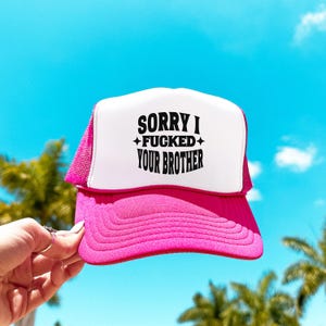May include: A bright pink and white trucker hat featuring a foam front panel with the black printed text SORRY I FUCKED YOUR BROTHER and small star accents. The hat has a pink mesh back, a curved pink brim, and a structured crown design.