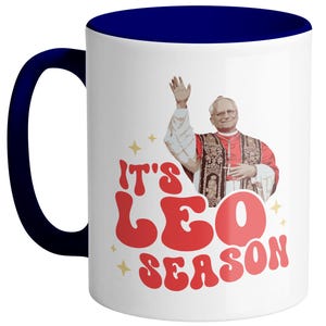 May include: White ceramic coffee mug with a blue rim. The mug features a retro-style graphic of a man in a red robe with the text "It's Leo Season".