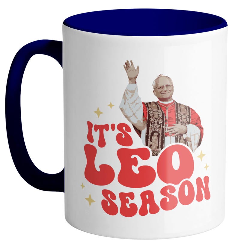May include: White ceramic coffee mug with a blue rim. The mug features a retro-style graphic of a man in a red robe with the text "It's Leo Season".