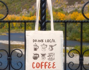 May include: Cream-colored tote bag with the words "DRINK LOCAL COFFEE" in black and red. The bag features illustrations of various coffee brewing methods. The background shows a river and trees with fall foliage.