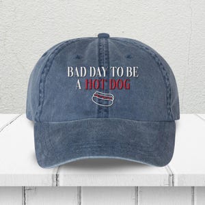 May include: A denim blue baseball cap with the embroidered phrase "BAD DAY TO BE A HOT DOG" in white and red. A graphic of a hot dog is below the text. The cap has a curved brim and a structured crown.