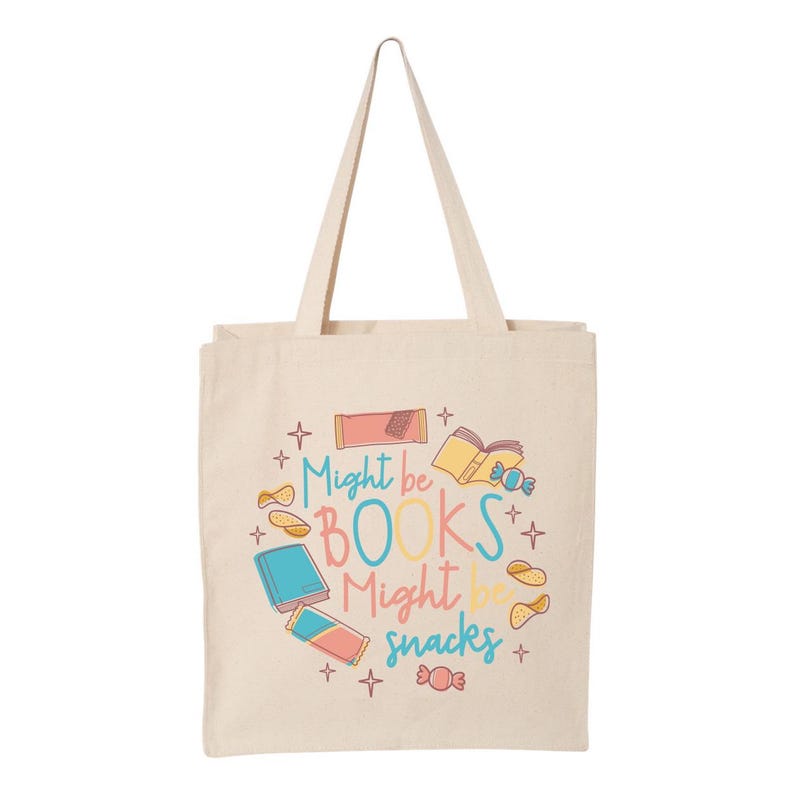 May include: A beige tote bag with a colourful design featuring the text "Might be BOOKS Might be snacks" and illustrations of books, snacks, and stars.