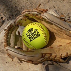 May include: A yellow softball with the text "Senior 2026" in black, resting in a well-worn, tan leather softball glove. The ball has red stitching. The glove is on a concrete surface.