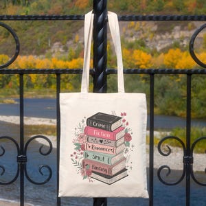 May include: Cream-colored tote bag featuring a stack of books with titles like "Crime," "Fiction," "Romance," "Smut," and "Fantasy." The books are decorated with floral and heart motifs. The bag has two long straps.