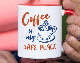 May include: White ceramic coffee mug with a red interior and handle. The mug features the text "Coffee is my SAFE PLACE" in orange and blue, along with a blue illustration of a person in a coffee cup.
