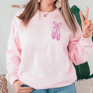 May include: A light pink sweatshirt featuring a graphic of two pink ballet shoes tied with a bow. The sweatshirt is a solid light pink color. The design is centered on the chest.