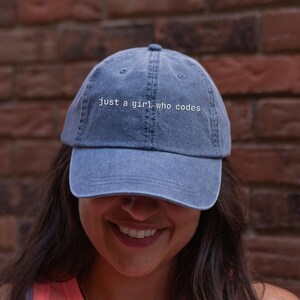 May include: A blue denim baseball cap with a curved brim and a button on top. The front of the cap features the white embroidered text "just a girl who codes" in a simple, lowercase sans-serif font. The cap has a six-panel construction.