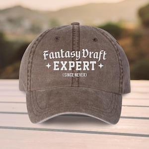 May include: A brown baseball cap with the text "Fantasy Draft EXPERT (SINCE NEVER)" in white script. The cap has a curved brim and a classic six-panel design, suitable for casual wear.
