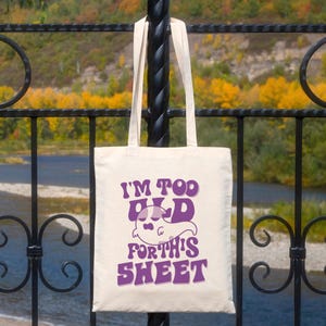 May include: A cream-colored tote bag with the text "I'M TOO OLD FOR THIS SHEET" in purple. The design includes a ghost illustration. The bag has two long handles and is hanging on a black metal fence.
