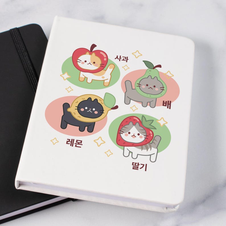 K&ouml;nnte beinhalten: Ein wei&szlig;es Notizbuch mit Illustrationen von Katzen in Obstkost&uuml;men: Apfel, Birne, Zitrone und Erdbeere. Jede Katze befindet sich in einem farbigen Oval mit koreanischem Text. Ein schwarzes Notizbuch ist teilweise sichtbar.