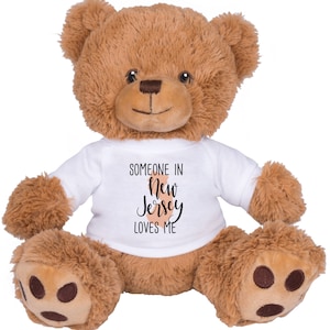 May include: A brown teddy bear wearing a white t-shirt with the text "SOMEONE IN New Jersey LOVES ME". The bear has a soft, plush texture and is sitting upright with its arms and legs extended.