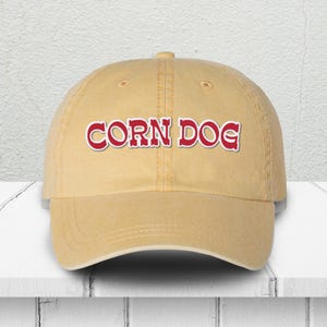 May include: A tan baseball cap with the words "CORN DOG" in red, outlined in white. The hat is made of a soft material and has a curved brim. The hat is sitting on a white surface.