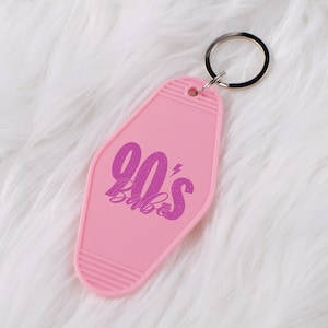 May include: A pink motel-style keychain with the text "90's Babe" in a purple, retro font. The keychain has a silver key ring and is set against a white, fluffy background.