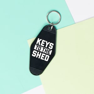 May include: Black motel key chain with the text "KEYS TO THE SHED" in white lettering.