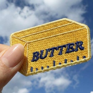 May include: Embroidered patch in the shape of a butter stick. The patch is yellow with the word "BUTTER" in navy blue. The patch is held against a blue sky with clouds.